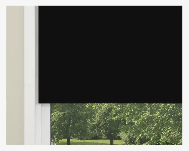Et nærbillede af et vindue med en sort rullegardin, der er delvist rullet ned. Gardinet er lavet af et uigennemsigtigt materiale, som effektivt blokerer lyset fra den øverste del af vinduet. Den nederste del af vinduet er ikke dækket, så man kan se ud til et grønt udendørsområde med træer og græs. Vinduesrammen er hvid, og væggen ved siden af er lysebeige.