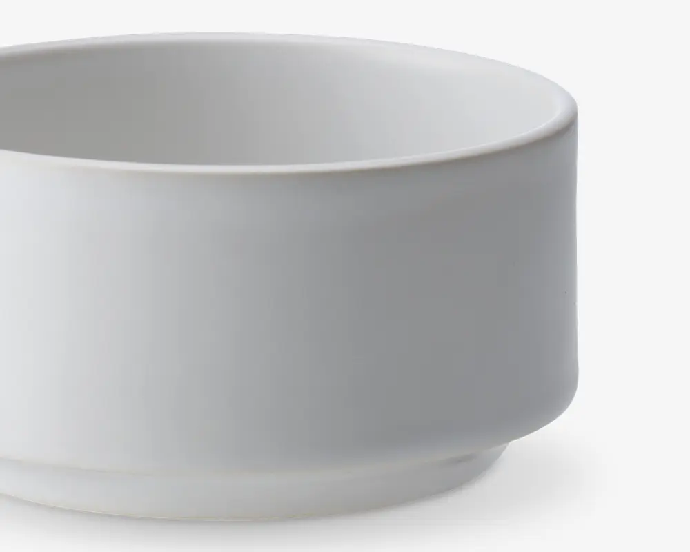 En enkel, hvid, cylindrisk skål med glatte sider og en let hævet bund. Skålen har et minimalistisk design uden mønstre eller dekorationer og en mat overflade.