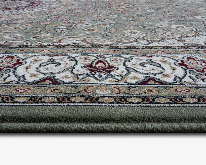 Nærbillede af et tykt, blødt tæppe med et traditionelt blomster- og snirklet mønster. Tæppet har en grøn bundfarve med indviklede designs i creme, beige, rød og sort. Kanten er dekoreret med detaljerede ornamenter, og luven ser tæt og blød ud, hvilket tyder på en behagelig tekstur at gå på.