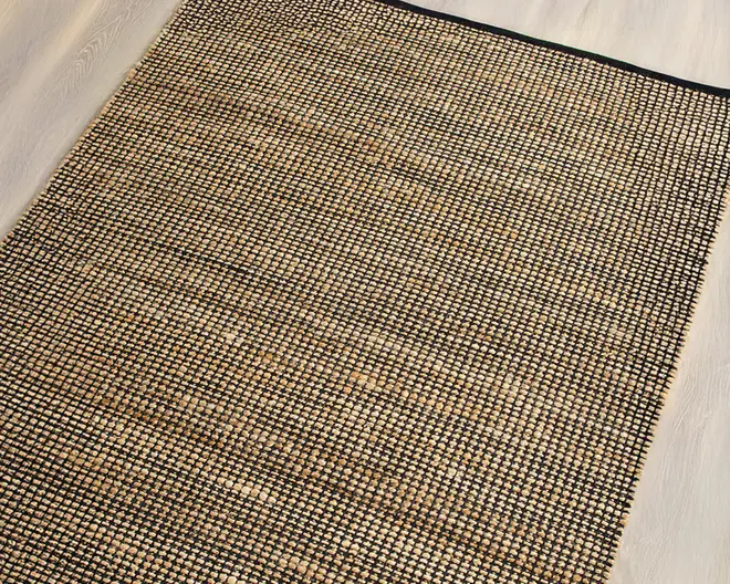 Rektangulært vævet tæppe med et gitterlignende mønster, der har naturlige beige og brune nuancer flettet sammen med sorte tråde. Kanterne er afsluttet med en solid sort kant for ekstra holdbarhed og et pænt udtryk. Teksturen virker grov og robust, velegnet til områder med meget trafik.