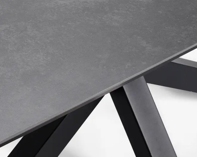 Nærbillede af et bordhjørne med en glat, mat grå overflade og en let stenlignende struktur. Bordpladen er tynd med rene, lige kanter. Benene er skråtstillede og lavet af solidt, mørkt metal, hvilket giver bordet et moderne og industrielt udtryk.