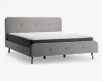 En moderne sengestel betrukket med lysegråt stof, med polstret hovedgærde og fodgærde, begge med enkle knapdetaljer. Sengen står på fire skrå træben i naturfinish, hvilket giver et retro-inspireret udtryk. Madrassen er tyk med hvid, quiltet top og mørkegrå sider.