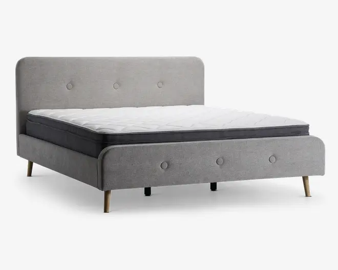 En moderne sengestel betrukket med lysegråt stof, med polstret hovedgærde og fodgærde, begge med enkle knapdetaljer. Sengen står på fire skrå træben i naturfinish, hvilket giver et retro-inspireret udtryk. Madrassen er tyk med hvid, quiltet top og mørkegrå sider.