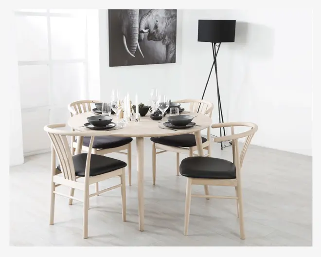 Et rundt spisebord i lyst træ med fire matchende stole, der har buede ryglæn og sorte polstrede sæder. Bordet er dækket med sorte tallerkener, skåle og glas, og pyntet med stearinlys og en lille dekoration i midten. Sættet står i et lyst, moderne rum med lyse gulve, en sort gulvlampe og et sort-hvidt elefantbillede på væggen.