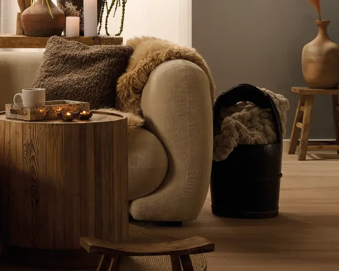 En hyggelig stue med en beige, buet sofa stylet med strukturerede brune puder og et faux fur-tæppe. Foran står et rundt sofabord i træ med en bakke, en kop og små lys, og ved siden af sofaen står en mat sort kurv med et kraftigt strikket tæppe. Varme trædetaljer og en neutral taburet med en vase fuldender udtrykket.