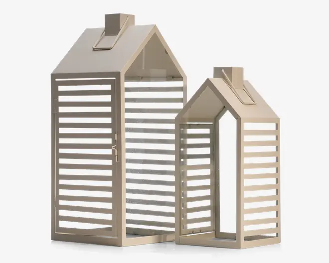 Sæt med to dekorative lanterner i form af små huse, fremstillet af lyst beige metal med lamel-design. Hver lanterne har saddeltag med en lille skorsten og en rektangulær bund. De åbne lameller på siderne lader lyset skinne igennem og skaber et mønstret lys. Lanternene kommer i to forskellige størrelser, én stor og én mindre, og har et moderne, minimalistisk udtryk.