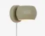 En moderne vægmonteret lampe med mat olivengrøn finish. Lampen har en rundet, let oval lampeskærm, der leder lyset nedad. Armaturet er fastgjort til en cirkulær vægplade, og en hvid ledning går ud fra bunden. Designet er minimalistisk og nutidigt, velegnet til forskellige indretninger.