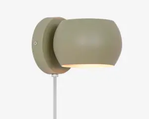 En moderne vægmonteret lampe med mat olivengrøn finish. Lampen har en rundet, let oval lampeskærm, der leder lyset nedad. Armaturet er fastgjort til en cirkulær vægplade, og en hvid ledning går ud fra bunden. Designet er minimalistisk og nutidigt, velegnet til forskellige indretninger.