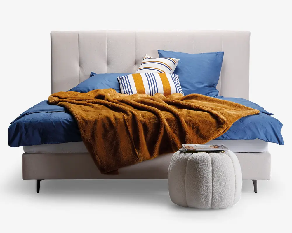 En moderne seng med en høj, polstret sengegavl i lys beige med knapper. Sengen er redt med dybblå lagner og pudebetræk, og der ligger et blødt, rustfarvet tæppe hen over sengen. Der er tre pyntepuder: én med blå og gule striber, én med blå og hvide striber og én ensfarvet blå. Foran sengen står en rund, hvid, tekstureret skammel med et magasin ovenpå.