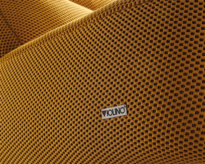 Nærbillede af et tekstureret stof i sennepsgul med et gitterlignende mønster, hvor der er fastgjort et lille rektangulært mærke med teksten 'VIOLINO'. Stoffet ser blødt ud og har en let hævet overflade, hvilket giver en taktil og visuelt interessant finish.