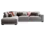 Stor L-formet hjørnesofa betrukket med lysegråt stof, med brede, dybe sæder og lav ryg. Sofaen er pyntet med flere pyntepuder i forskellige teksturer og farver, herunder brune, rustfarvede og lyserøde nuancer, hvilket giver både komfort og et dekorativt udtryk. Venstre side har en udvidet chaiselong for ekstra afslapning.