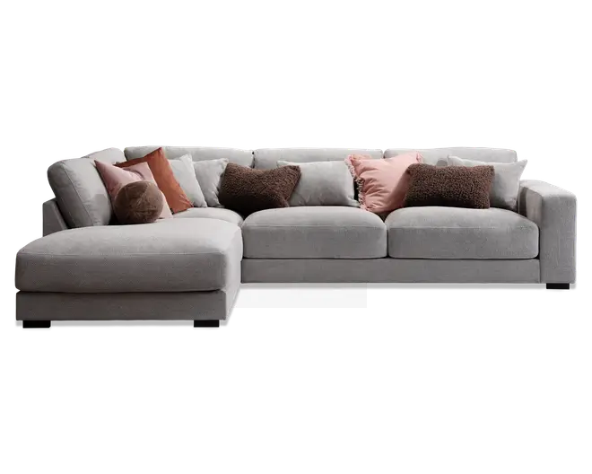 Stor L-formet hjørnesofa betrukket med lysegråt stof, med brede, dybe sæder og lav ryg. Sofaen er pyntet med flere pyntepuder i forskellige teksturer og farver, herunder brune, rustfarvede og lyserøde nuancer, hvilket giver både komfort og et dekorativt udtryk. Venstre side har en udvidet chaiselong for ekstra afslapning.