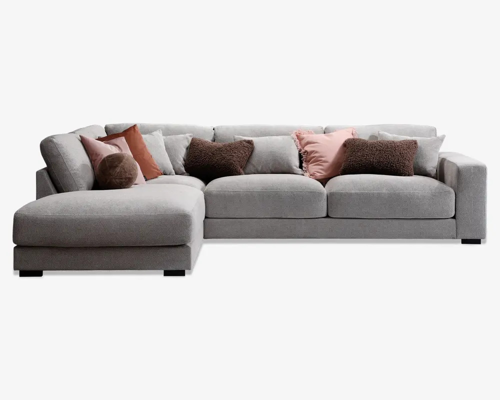 Stor L-formet hjørnesofa betrukket med lysegråt stof, med brede, dybe sæder og lav ryg. Sofaen er pyntet med flere pyntepuder i forskellige teksturer og farver, herunder brune, rustfarvede og lyserøde nuancer, hvilket giver både komfort og et dekorativt udtryk. Venstre side har en udvidet chaiselong for ekstra afslapning.