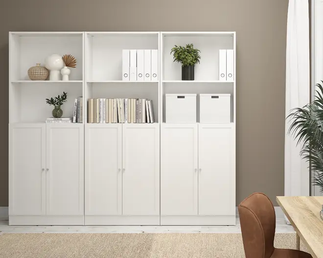 Høj, hvid reolenhed med tre sektioner, hver med to lukkede skabe nederst og åbne hylder øverst. Hylderne indeholder en blanding af dekorative genstande som vaser og en plante, samt bøger, hvide opbevaringskasser og hvide ringbind. Designet er minimalistisk og moderne med rene linjer og mat finish.