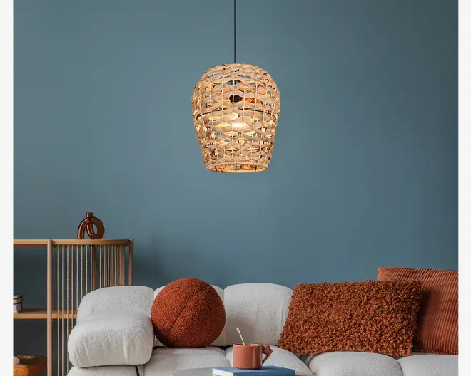 Hængelampe med en skærm flettet af rattan i en naturlig beige farve, med et åbent gitterdesign, der lader lyset skinne igennem og skaber en varm, tekstureret glød. Skærmen har en rundet, let tilspidset form og hænger i en tynd sort ledning. Udtrykket er afslappet og moderne, velegnet til hyggelige eller boheme-inspirerede indretninger.