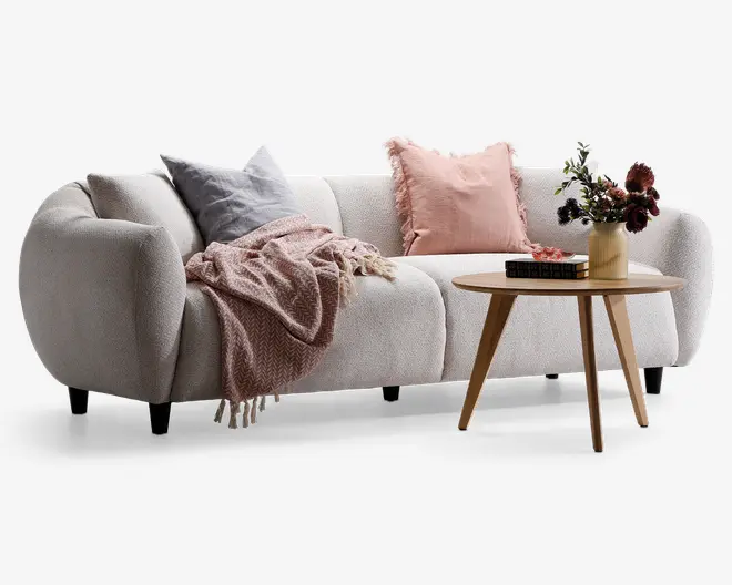 En moderne, blød sofa med afrundede armlæn og et blødt, tekstureret betræk i lys beige. Sofaen er pyntet med to pyntepuder – én i afdæmpet grå og én i støvet rosa med frynset kant. Et hyggeligt, mønstret tæppe i støvet rosa er lagt over den ene side. Foran sofaen står et rundt sofabord i lyst træ med en vase med mørke og lyse blomster samt et par stablede bøger, hvilket giver indretningen et elegant og komfortabelt udtryk.