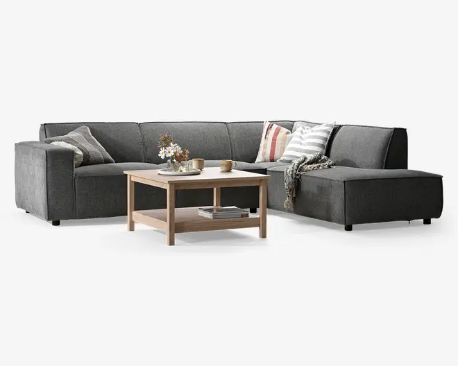 En moderne L-formet hjørnesofa betrukket med mørkegråt stof, med brede armlæn og lav ryg. Sofaen er pyntet med flere dekorative puder i forskellige mønstre og farver samt et hyggeligt tæppe lagt over den ene side. Foran sofaen står et sofabord i lyst træ med en underhylde, hvor der er en blomsterdekoration, kopper og bøger, hvilket giver både stil og funktionalitet.