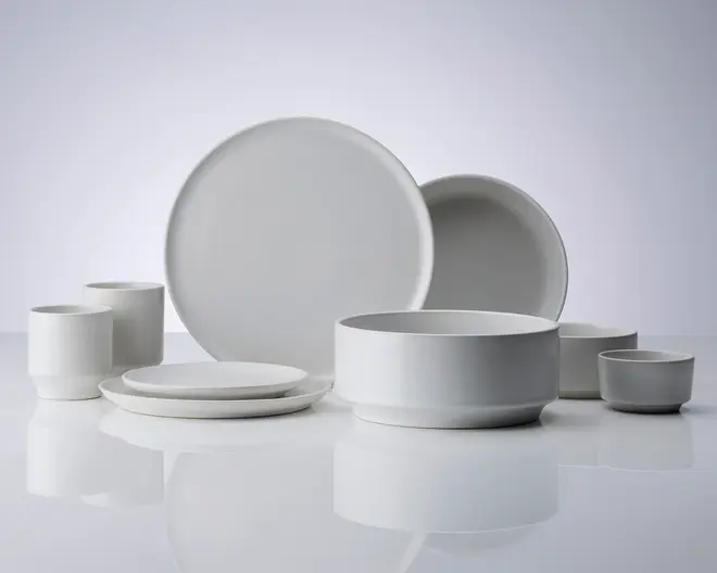 Et sæt minimalistisk, mat hvidt keramikstel bestående af to store middagstallerkener, to mindre sidetallerkener, to cylindriske krus, en stor serveringsskål og to små skåle. Designet er moderne med rene linjer og glatte overflader, hvilket giver et stilrent og elegant udtryk. Delene kan stables og har en ensartet farve uden mønstre eller dekorationer.