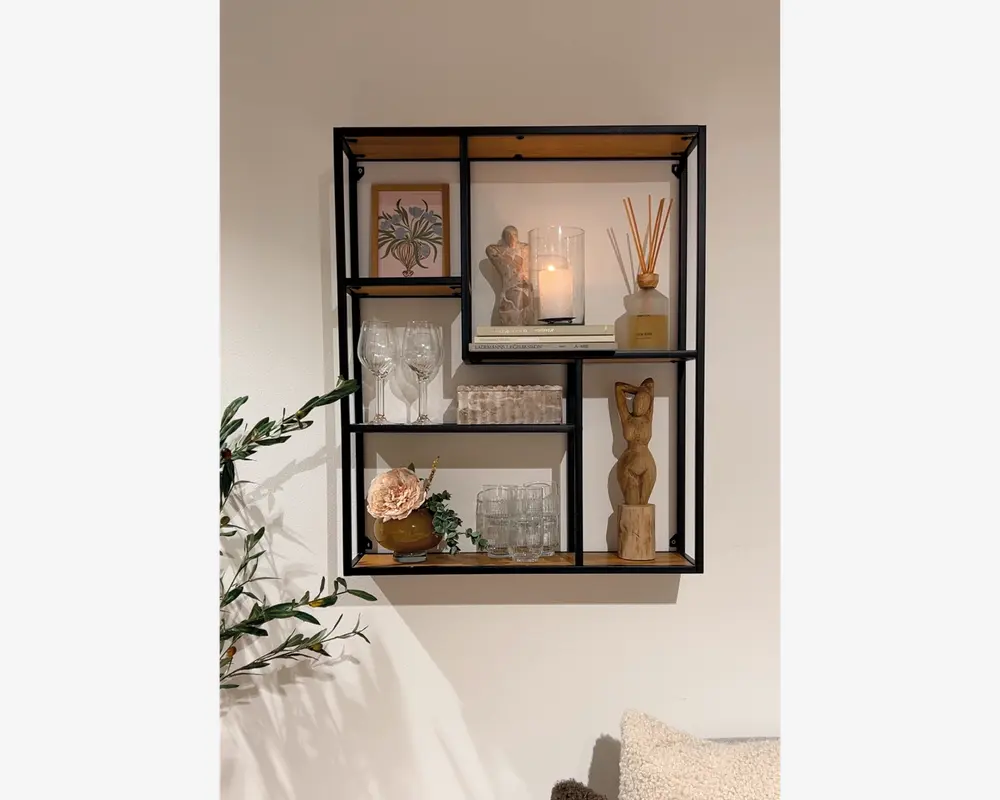 Vægmonteret reol med sort metalramme og træhylder, designet i et moderne geometrisk mønster med flere rum. Hylderne er dekoreret med forskellige pyntegenstande, herunder et indrammet botanisk billede, en glascylinder med tændt lys, en duftpind-diffuser, en lille træskulptur, vinglas, drikkeglas, en kunstig blomsterdekoration i en lille vase og en stak bordskånere. Udtrykket er moderne og stilfuldt, velegnet til stue eller entré.