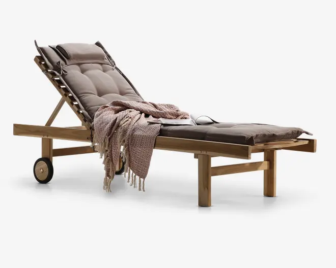 Justerbar liggestol i træ med tykke, polstrede brune hynder for ekstra komfort. Stolen har en tilbagelænet ryg, to hjul for nem flytning og en solid ramme. Et tekstureret, frynset tæppe i støvet rosa farve er lagt over sædet, og et par solbriller ligger på hynden, hvilket giver et indtryk af afslapning udendørs.
