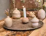 En dekorativ opstilling med små, runde keramikvaser i bløde beige og cremefarver, nogle med fine kunstige blomster. En vase i midten fungerer som lysestage med et tændt hvidt kronelys. Elementerne er placeret på et stort, tekstureret metalfad oven på et rustikt træbord, hvilket skaber et hyggeligt og elegant udtryk.