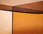 Nærbillede af et møbel med tonede glasplader i en varm ravfarvet nuance. Glasset har en glat, blank overflade og skarpe, rene kanter, der mødes i en ret vinkel. Glassets gennemsigtighed og farve giver et moderne, elegant udtryk med overlappende refleksioner og en gradient-effekt.