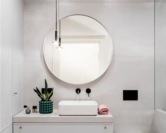 Moderne badeværelsesindretning med et stort rundt spejl monteret på en lysegrå flisevæg. Bordpladen er hvid med en rektangulær fritstående vask og mat sorte vægmonterede armaturer. Dekorationerne omfatter en grøn vase med slangplante, et lille stearinlys, en sort beholder og et pænt sammenrullet lyserødt håndklæde. To minimalistiske pendellamper hænger fra loftet og giver et moderne udtryk.
