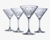 Sæt med fire klare glas med stilk, hver med en bred, konisk skål og indviklede geometriske mønstre skåret i den nederste del af skålen. Glassene har en solid fod og elegante, lange stilke, hvilket giver dem et sofistikeret og klassisk udseende.