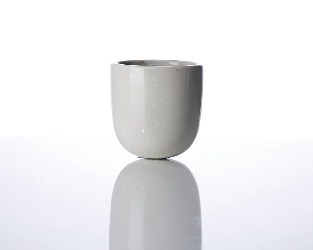 En lille, rund, lysegrå keramikkop med en glat mat overflade. Koppen har lige sider og en let afrundet bund, uden synligt håndtag eller dekoration. Det minimalistiske design giver den et moderne og elegant udtryk.