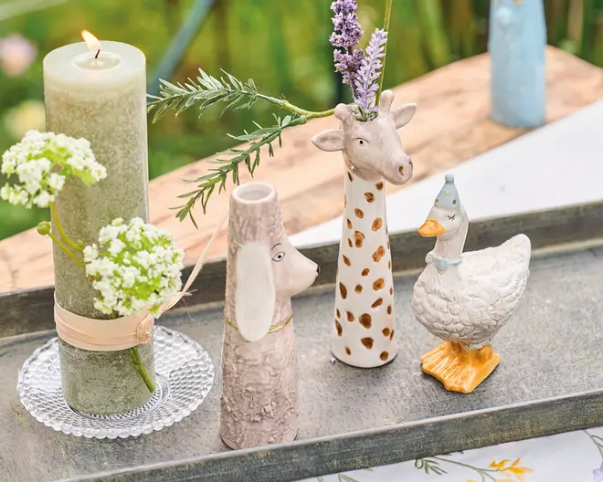 Billedet viser en dekorativ opstilling med et højt grønt stearinlys, der har en blomsterdekoration bundet omkring sig og står på en glasunderkop. Ved siden af lyset er der tre keramiske dyrefigurer: en vase formet som et hundehoved og hals med tekstureret pels, en girafvase med lang hals, brune pletter og lavendelstilke i, samt en hvid gåsefigur med blå prikket hat og orange fødder. Alle genstandene står på et rektangulært metalfad, placeret på et bord udendørs.