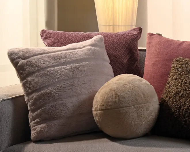 Udvalg af dekorative pyntepuder på en grå sofa, herunder en stor blød grå firkantet pude, en mauve-mønstret pude, en pink pude, en struktureret mørkebrun pude og en rund beige pude.