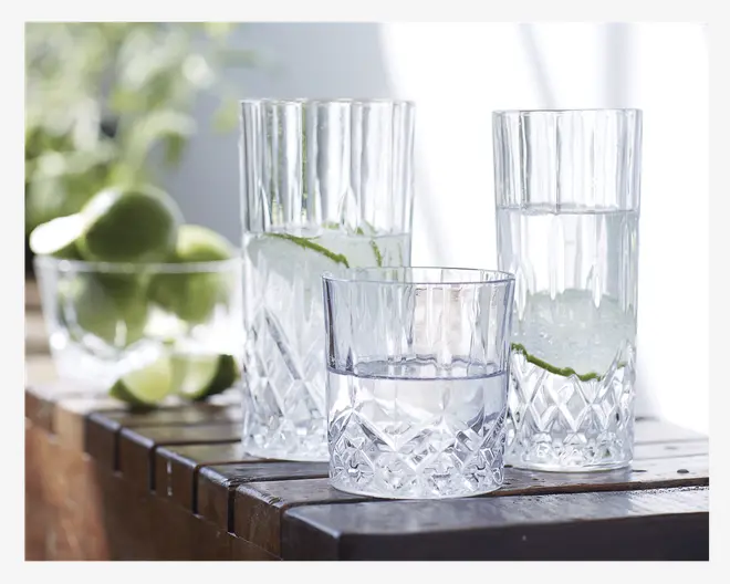 Tre klare drikkeglas med facetslebet diamantmønster i bunden og lodrette riller langs siderne. To af glassene er høje og cylindriske, mens det tredje er lavere og bredere. Glassene er fyldt med vand og pyntet med skiver af lime. Glassene står på en træoverflade med en skål friske limefrugter i baggrunden.