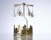 Et dekorativt ornament i metal med en rund base, der er udsmykket med udskårne grantræer. Over basen er der en lodret stang, som bærer et roterende karrusel med hængende vedhæng formet som juletræer. Hele ornamentet er lavet af blankt guldfarvet metal, hvilket giver et festligt og elegant udtryk. Karrusellen er designet til at dreje rundt, sandsynligvis drevet af varme fra stearinlys placeret i bunden (ikke vist på billedet).