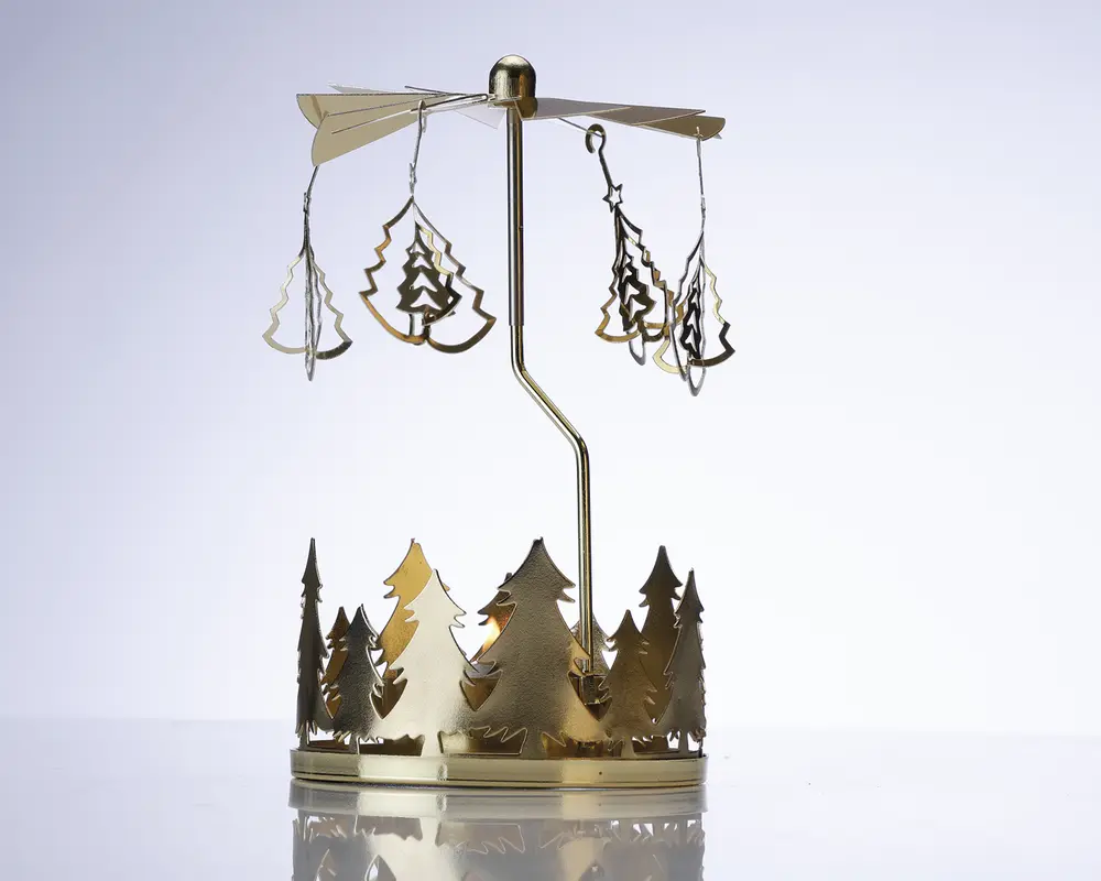 Et dekorativt ornament i metal med en rund base, der er udsmykket med udskårne grantræer. Over basen er der en lodret stang, som bærer et roterende karrusel med hængende vedhæng formet som juletræer. Hele ornamentet er lavet af blankt guldfarvet metal, hvilket giver et festligt og elegant udtryk. Karrusellen er designet til at dreje rundt, sandsynligvis drevet af varme fra stearinlys placeret i bunden (ikke vist på billedet).