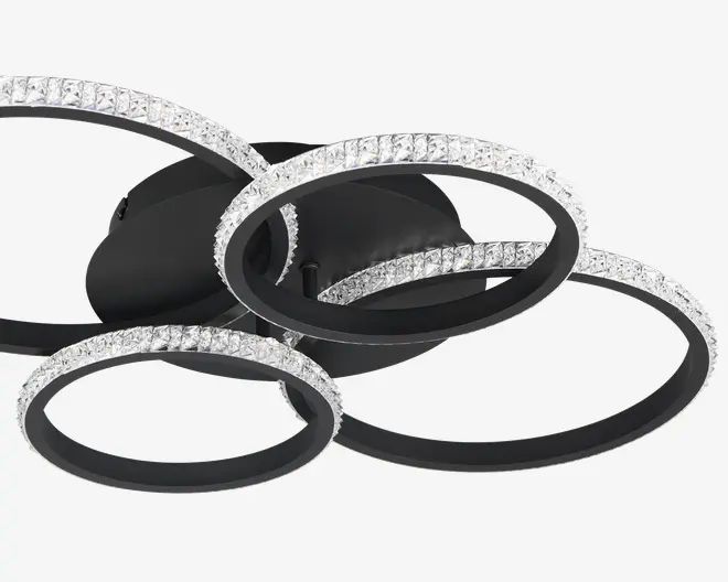 Loftlampe med fire sammenflettede sorte metalringe, hver dekoreret med en kant af klare, facetslebne krystaller, der forstærker lysets refleksion. Ringene er arrangeret i et moderne, overlappende design, hvilket giver et markant geometrisk udtryk. Krystallerne er placeret i en ubrudt linje langs ydersiden af hver ring, hvilket tilføjer et luksuriøst og elegant præg.