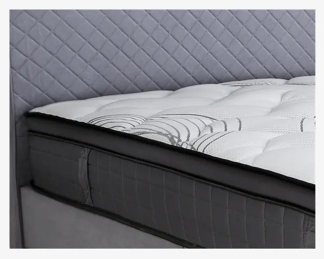 Nærbillede af et madras-hjørne med en tyk, blød, quiltet top i hvidt stof med diskrete sorte swirl-mønstre. Siderne er betrukket med mørkegråt stof med kvadratsyninger, og madrassen har forstærkede kanter for ekstra holdbarhed.