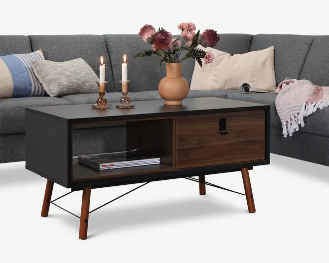 Rektangulært sofabord med mat sort ramme og skråtstillede træben. Bordet har en åben hylde på den ene side til opbevaring, hvor der ligger magasiner, og en enkelt skuffe med trælook og sort greb på den anden side. Bordpladen er pyntet med en keramikvase med lyserøde blomster og to trælysestager med tændte stearinlys.