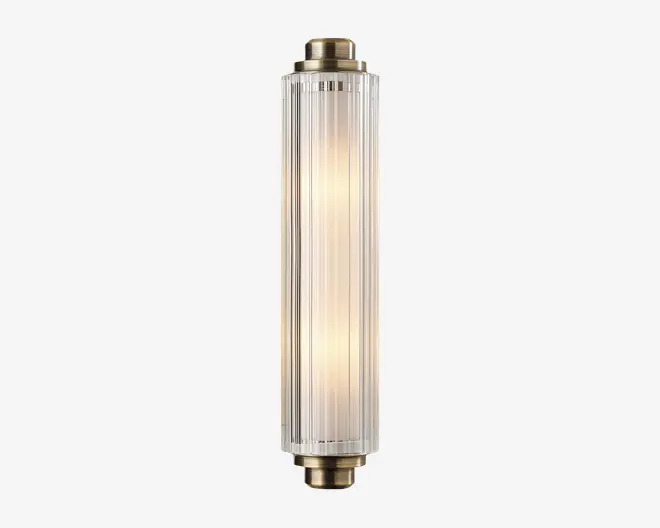 En lodret vægmonteret lampe med cylindrisk form og klare, riflede glaspaneler. Enderne er afsluttet med metalbeslag i messingfarve, hvilket giver lampen et vintage-inspireret udtryk. Lyset indeni udsender en varm, diffus glød gennem det strukturerede glas, hvilket skaber et elegant og sofistikeret udseende.
