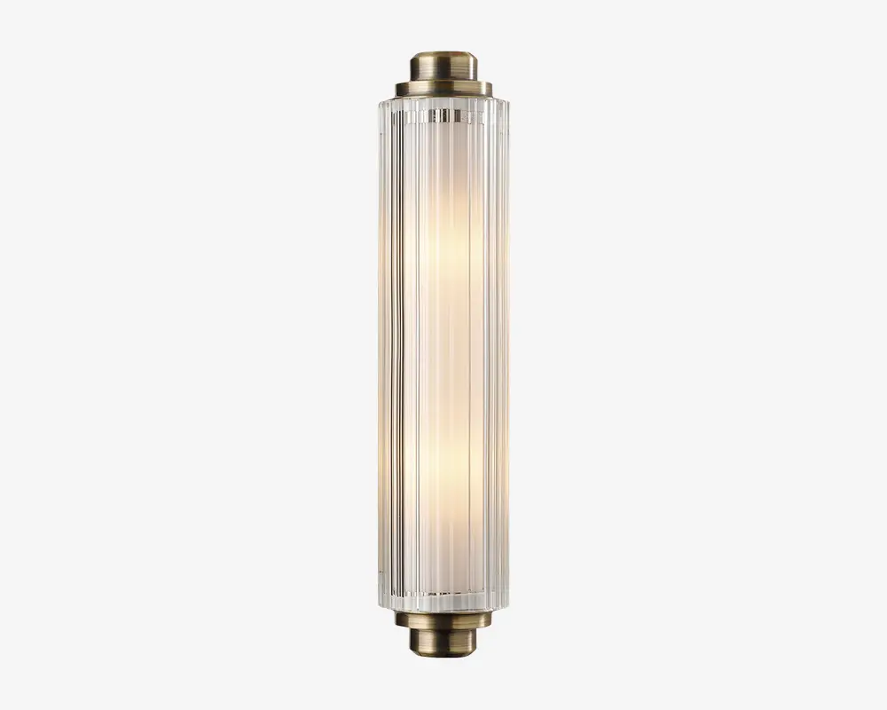 En lodret vægmonteret lampe med cylindrisk form og klare, riflede glaspaneler. Enderne er afsluttet med metalbeslag i messingfarve, hvilket giver lampen et vintage-inspireret udtryk. Lyset indeni udsender en varm, diffus glød gennem det strukturerede glas, hvilket skaber et elegant og sofistikeret udseende.