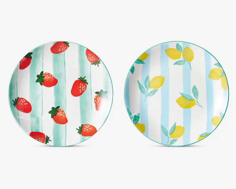 To runde tallerkener med farverige frugtmotiver. Den venstre tallerken har et mønster af røde jordbær med grønne blade på en hvid baggrund med lodrette grønne akvarelstriber. Den højre tallerken viser gule citroner med grønne blade på en hvid baggrund med lodrette blå akvarelstriber. Begge tallerkener har en blank overflade og et legende, sommerligt udtryk.