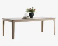 Rektangulært bord med lys træfinish, rene linjer og let tilspidsede ben. Bordpladen er glat og rummelig, dekoreret med en mørkegrøn vase med lyserøde og hvide blomster, en stak tallerkener og to hvide stearinlys placeret på flettede dækkeservietter.