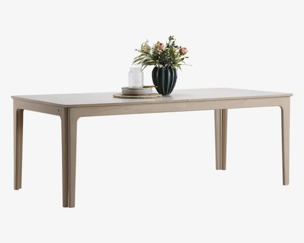 Rektangulært bord med lys træfinish, rene linjer og let tilspidsede ben. Bordpladen er glat og rummelig, dekoreret med en mørkegrøn vase med lyserøde og hvide blomster, en stak tallerkener og to hvide stearinlys placeret på flettede dækkeservietter.