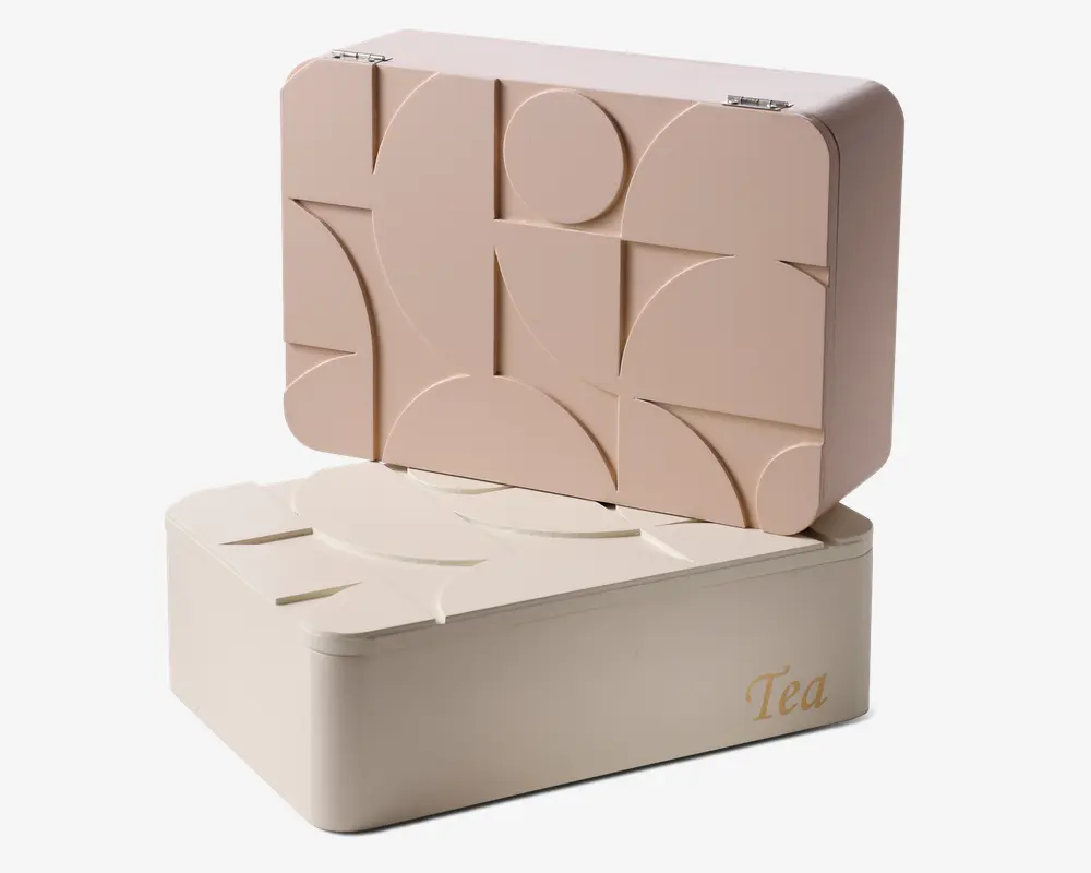 Rektangulær opbevaringsboks med hængslet låg, der har et hævet geometrisk mønster af cirkler og linjer på ydersiden. Boksen er i en blød beige farve med et lidt mørkere låg, og ordet 'Tea' er elegant trykt med guldfarvet skrift på fronten. Designet er moderne og minimalistisk med glatte, afrundede kanter.