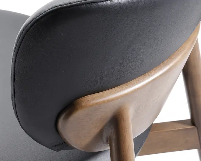 Nærbillede af en stol med en glat, sort polstret ryg med en let tekstur. Ryggen understøttes af en buet, lysebrun træramme, der forbinder til afrundede træben og fremhæver kombinationen af blødt polster og naturlige træmaterialer.