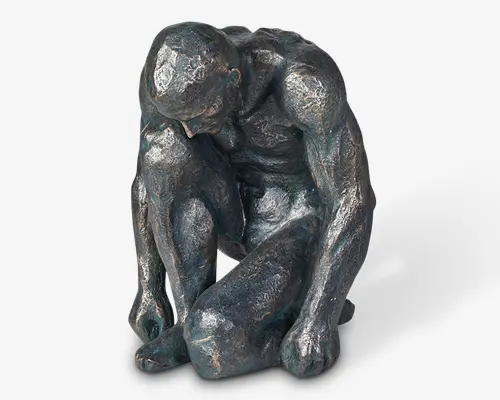 En lille, detaljeret skulptur af en muskuløs mandlig figur i en sammenkrøbet, eftertænksom positur med hovedet bøjet og armene hvilende på bøjede knæ. Overfladen har en tekstureret, stenlignende finish med en mørk, metallisk patina, hvilket giver den et antikt og kunstnerisk udtryk. Figurens anatomi er veldefineret og fremhæver musklernes og kroppens konturer.