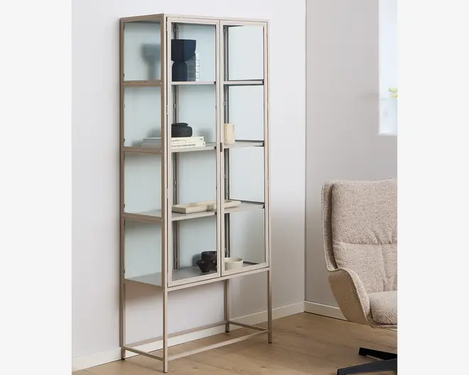 Høj, rektangulær vitrineskab med let metalramme og glaslåger. Skabet har fire glashylder og matterede glas sidepaneler, hvilket giver et moderne og luftigt udtryk. Lågerne har smalle metalhåndtag, og skabet står på slanke ben, der løfter det fra gulvet. Indeni er forskellige dekorative genstande arrangeret, hvilket viser hyldernes rummelighed og alsidighed.