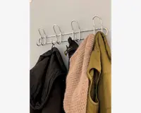 Væghængt knagerække i metal med seks store, buede kroge. Krogene er jævnt fordelt og fastgjort til en vandret metalstang. På knagerækken hænger en sort jakke, en beige strikket sweater, en grøn jakke og et par sorte handsker. Designet er minimalistisk og funktionelt, velegnet til organisering af overtøj og tilbehør.