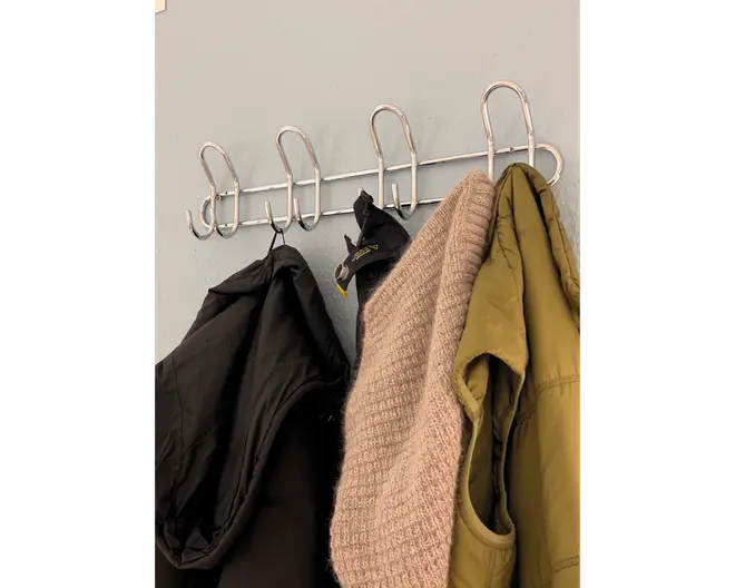 Væghængt knagerække i metal med seks store, buede kroge. Krogene er jævnt fordelt og fastgjort til en vandret metalstang. På knagerækken hænger en sort jakke, en beige strikket sweater, en grøn jakke og et par sorte handsker. Designet er minimalistisk og funktionelt, velegnet til organisering af overtøj og tilbehør.