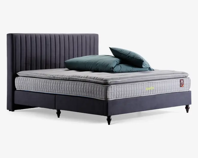 En moderne seng med en høj, lodret-stiksyet sengegavl i mørkegråt stof. Madrassen er tyk og har et quiltet, ternet lysegråt betræk med en blød topmadras. Sengebunden er polstret i samme mørkegrå stof og står på korte, sorte ben. To teal-farvede puder og en matchende dyne ligger pænt oven på madrassen.