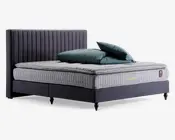 En moderne seng med en høj, lodret-stiksyet sengegavl i mørkegråt stof. Madrassen er tyk og har et quiltet, ternet lysegråt betræk med en blød topmadras. Sengebunden er polstret i samme mørkegrå stof og står på korte, sorte ben. To teal-farvede puder og en matchende dyne ligger pænt oven på madrassen.